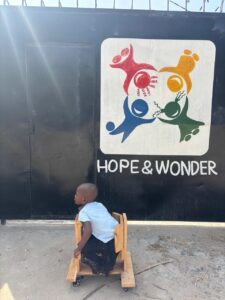 Kind aus Afrika sitzt auf dem Holzwägelchen vor einem Plakat "Hope & Wonder"